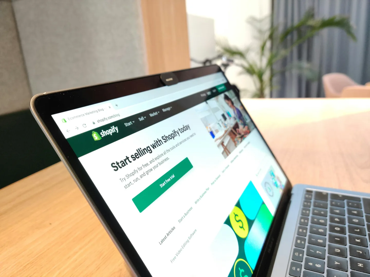 Laptop on a wooden desk displaying the Shopify homepage with a 'Start selling with Shopify today' headline and a green call-to-action button.
